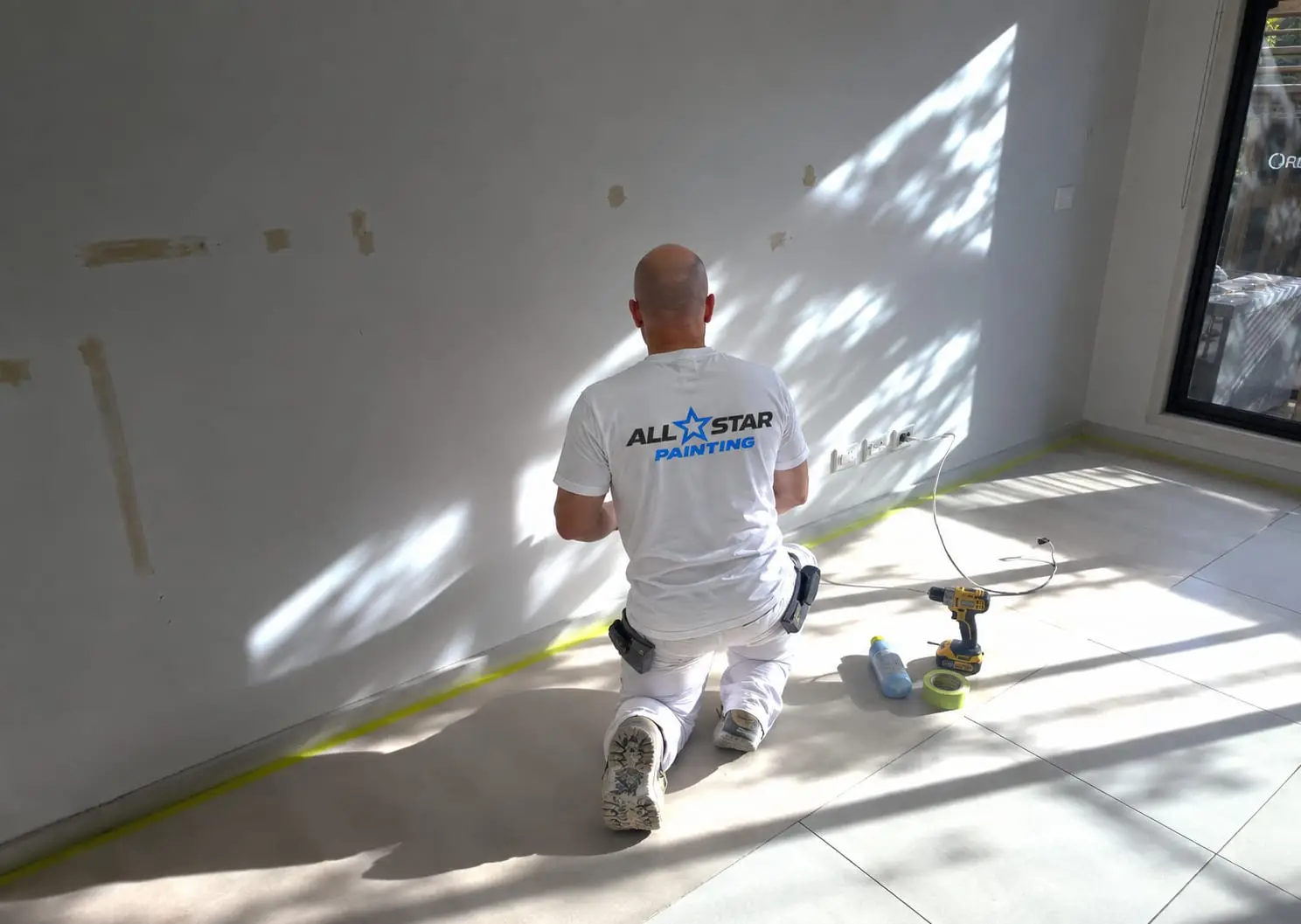 All Star painter kneeling to prepare a filled wall, skirting masked in yellow tape